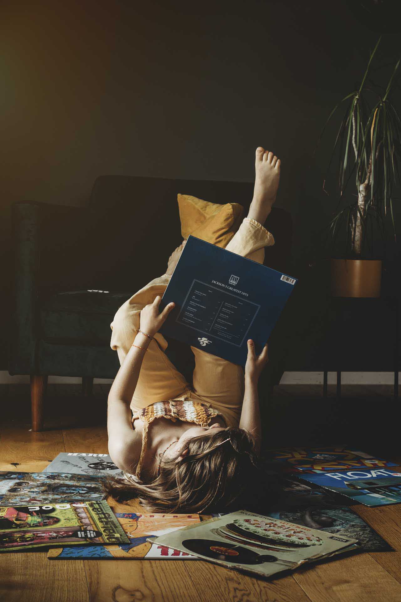 Person lying on the floor reading a vinyl record sleeve titled “Jackson’s Greatest Hits,” surrounded by scattered records and magazines beside a dark armchair.