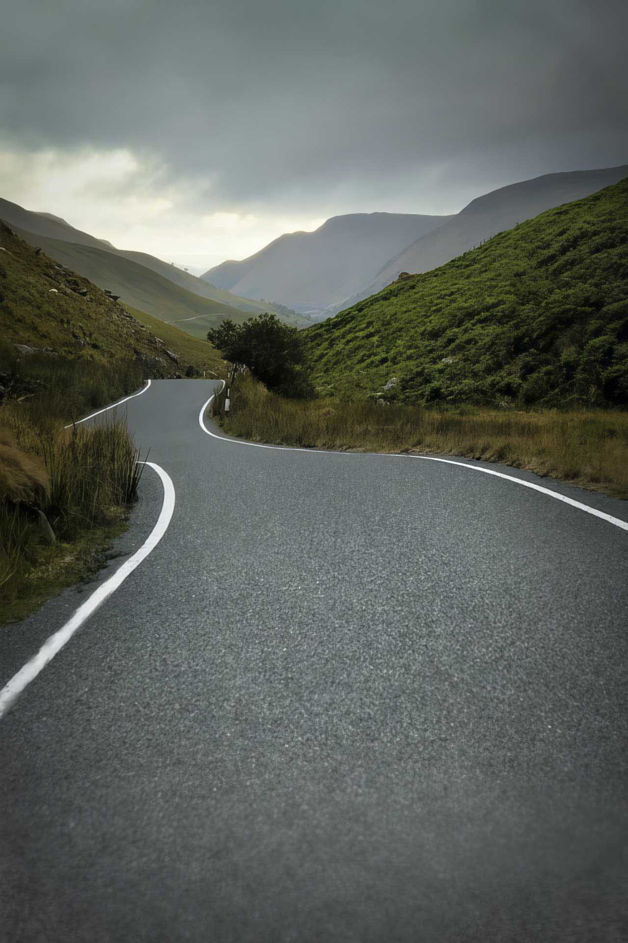 Winding mountain road curving through green hills beneath heavy grey clouds, leading into a distant misty valley.