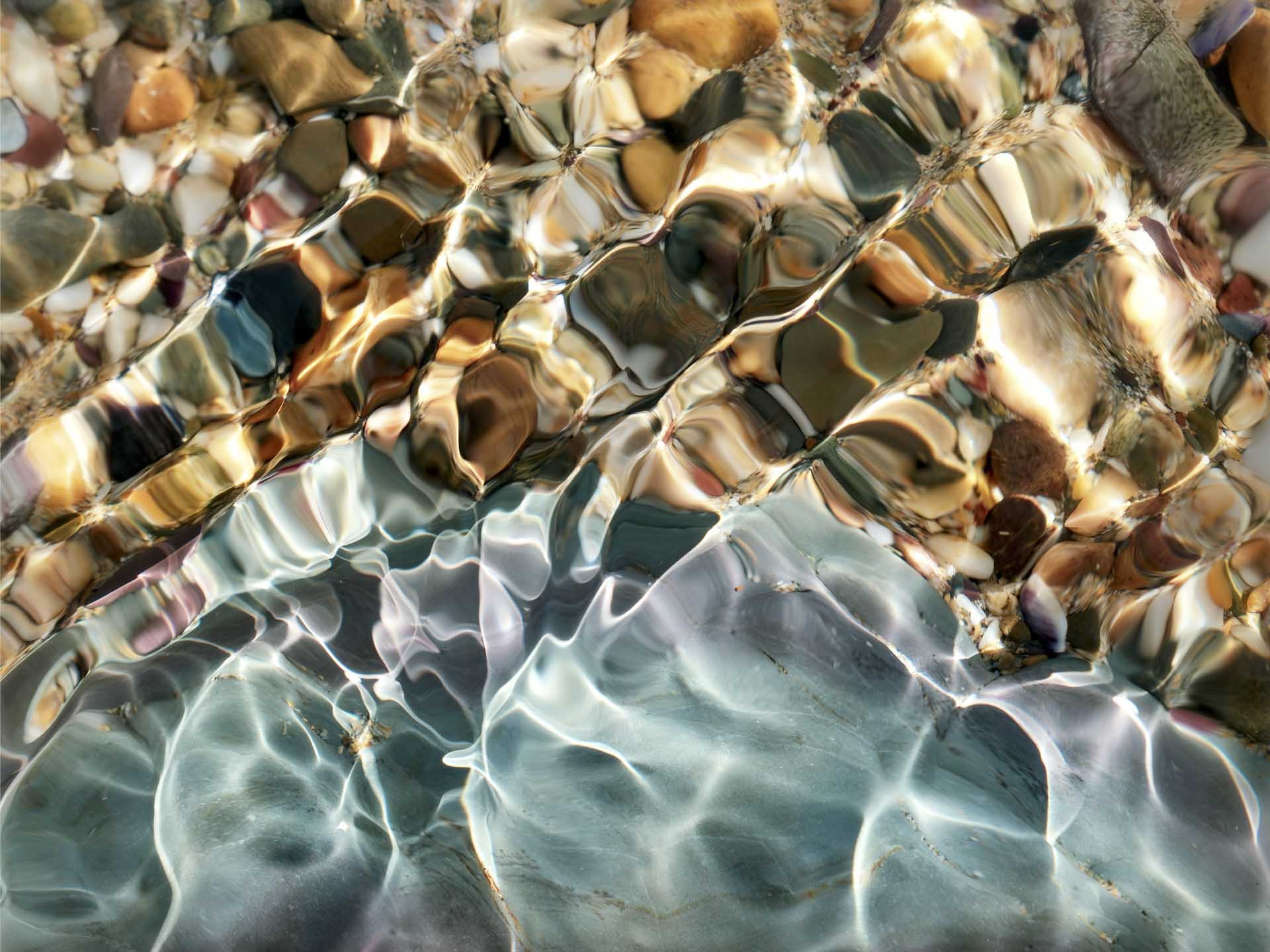Close-up of clear water rippling over multicoloured pebbles, sunlight creating bright shifting patterns on the stones.