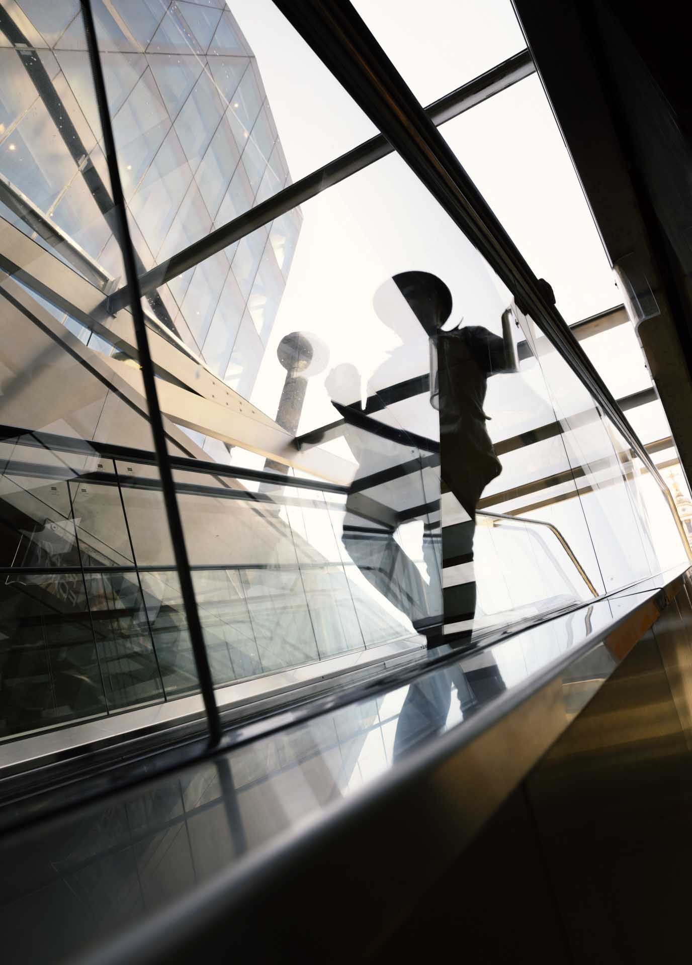 Silhouette of a person riding an escalator inside a glass structure with reflections and a modern building visible outside.