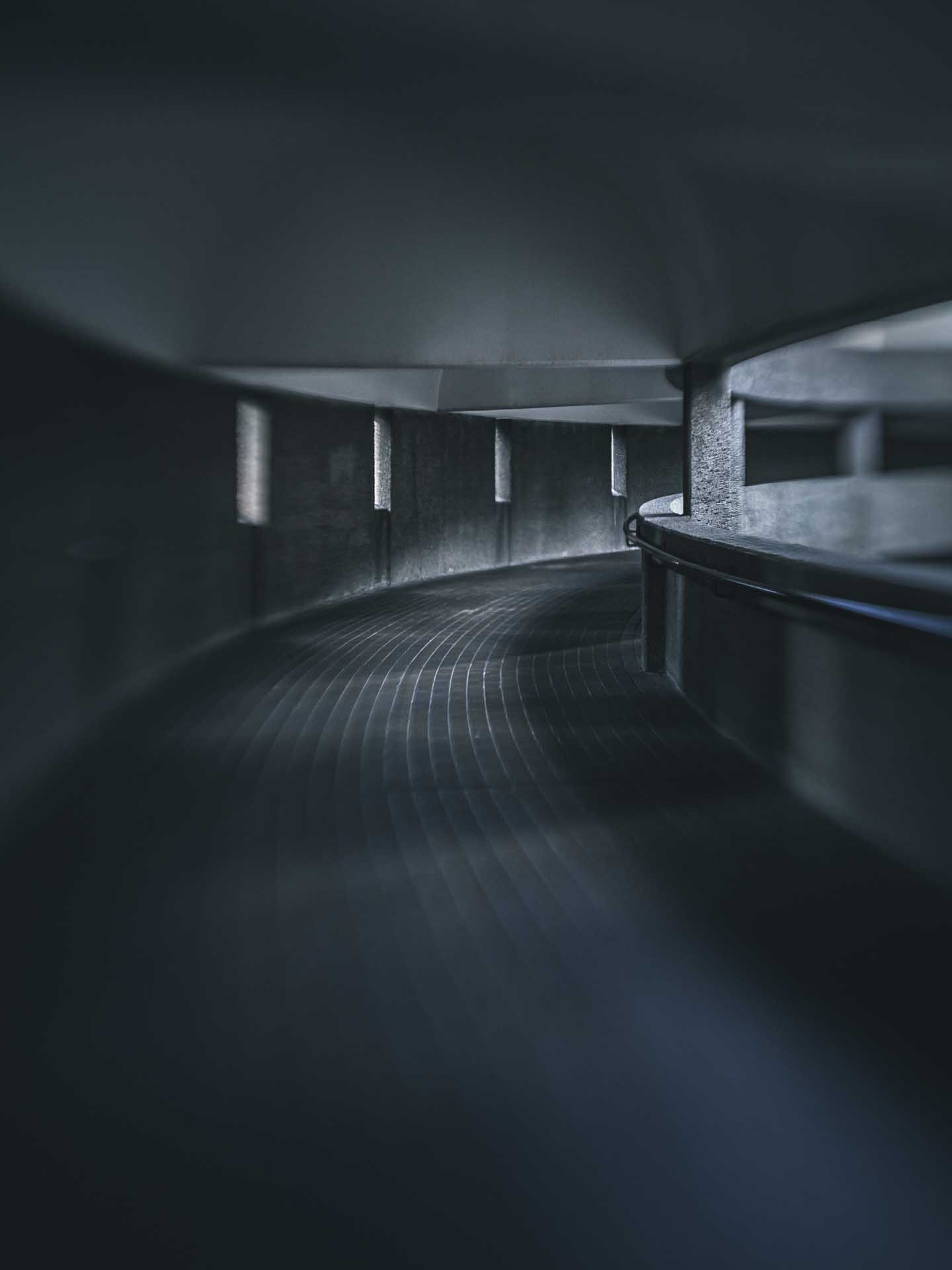 Dimly lit curved concrete corridor with a tiled floor and small rectangular wall lights receding into the distance.