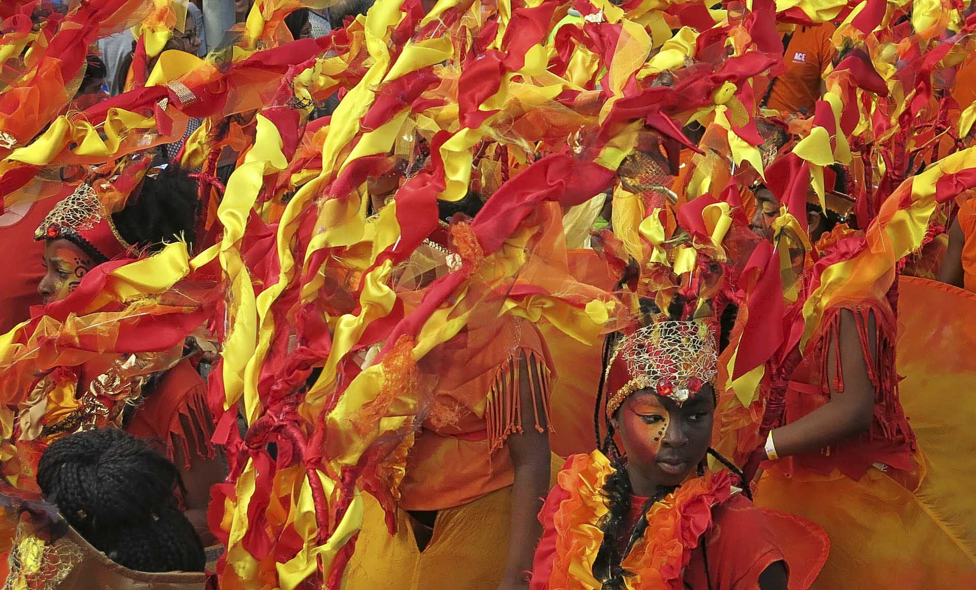 Crowd of carnival performers wearing elaborate red and yellow costumes with ribbons and face paint.