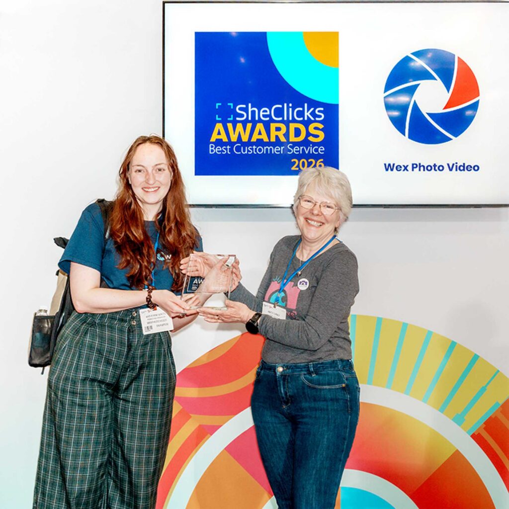 Two women smiling while holding a glass award in front of a screen reading “SheClicks Awards Best Customer Service 2026” with Wex Photo Video logo.