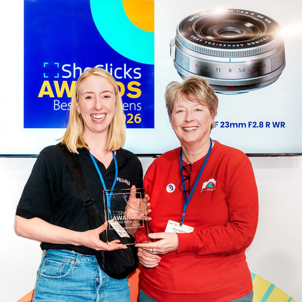 Two women smiling and presenting an award; screen shows “SheClicks Awards Best Prime Lens 2026” with a Fujifilm 23mm lens image.