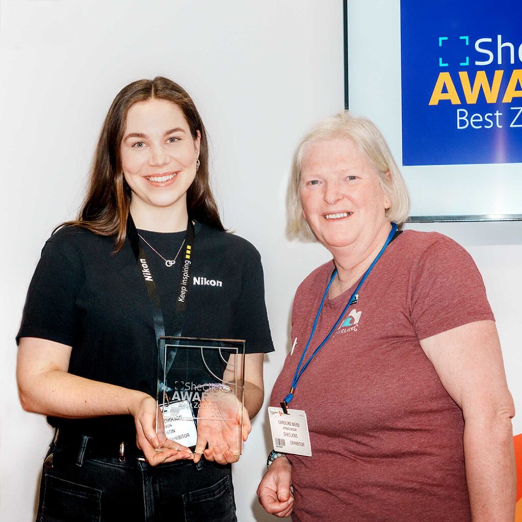 Two women holding an award; one wears a Nikon lanyard. Screen reads “SheClicks Awards Zoom Lens 2026.”