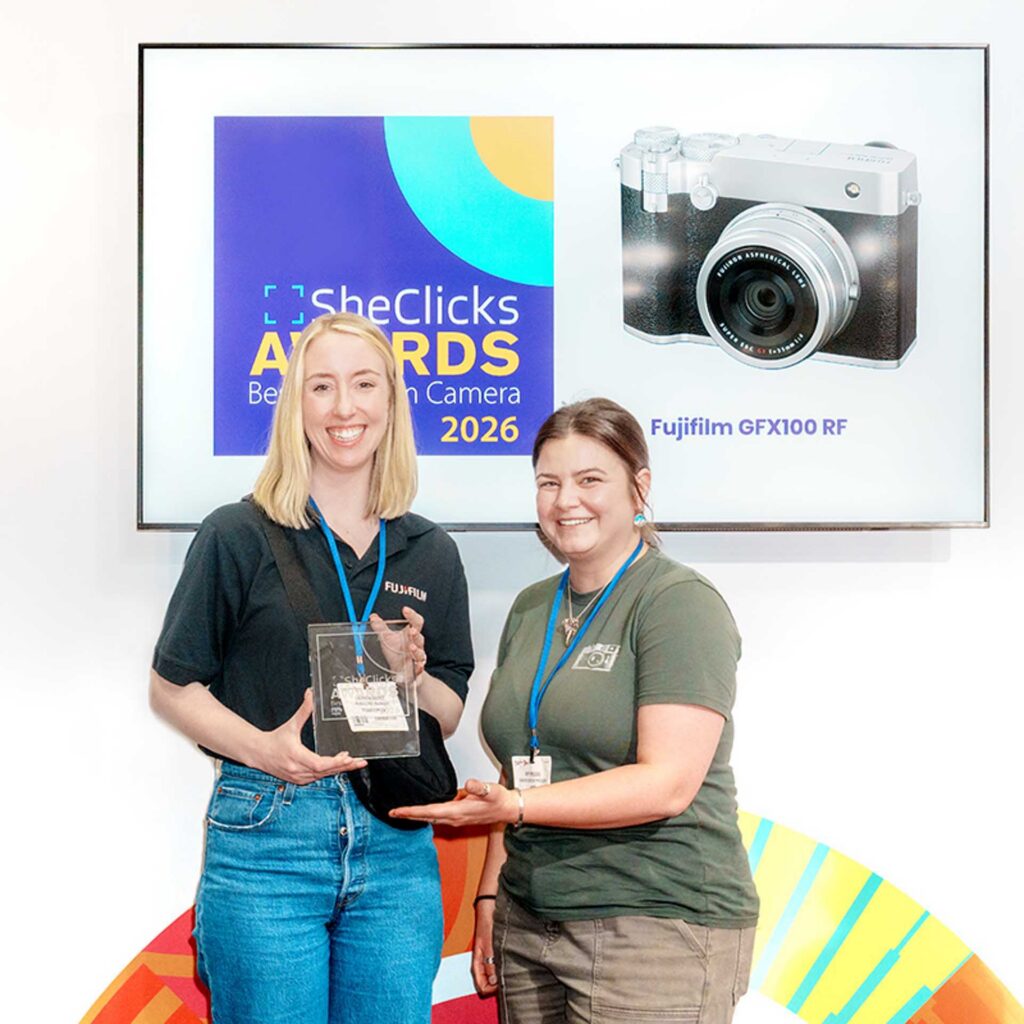 Two women smiling with an award; screen behind shows “Fujifilm GFX100 RF” and “SheClicks Awards Best Camera 2026.”