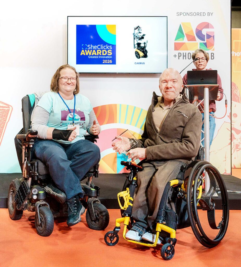 Two wheelchair users holding an award on stage; screen reads “SheClicks Awards Greatest Innovation 2026” with sponsor signage visible.