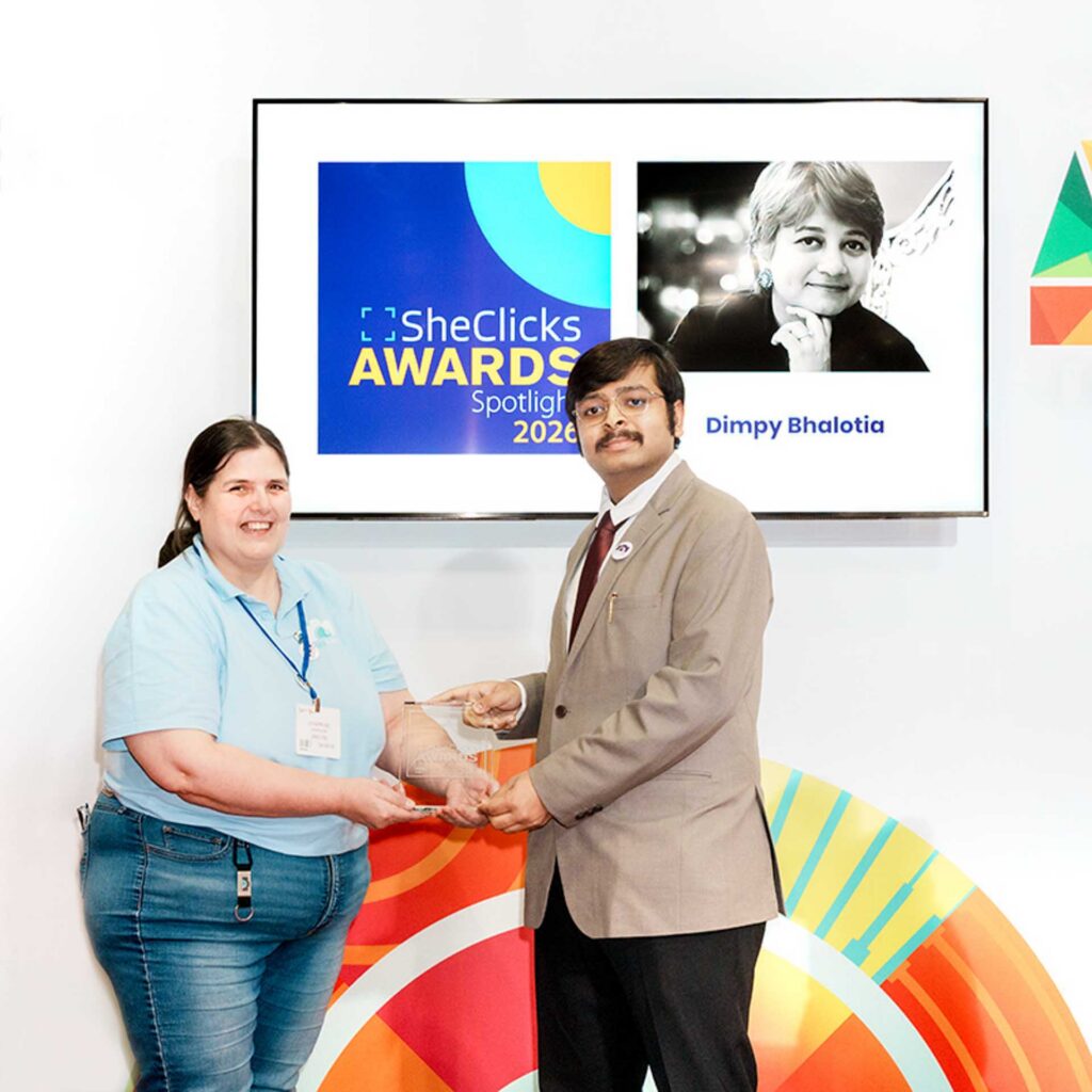 Woman and man in suit holding an award; screen behind reads “SheClicks Awards Spotlight 2026” and “Dimpy Bhalotia.”