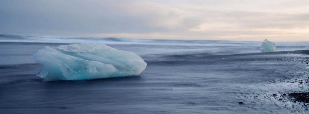 Large pale-blue iceberg resting on a black-sand beach, with waves blurred by a long exposure under a gray sky.