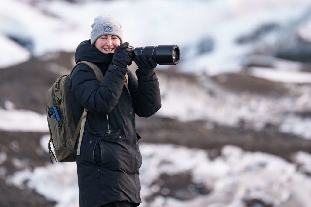 Smiling photographer (Emily Endean) in winter clothing holding a camera with a telephoto lens, standing in a snowy Icelandic landscape.