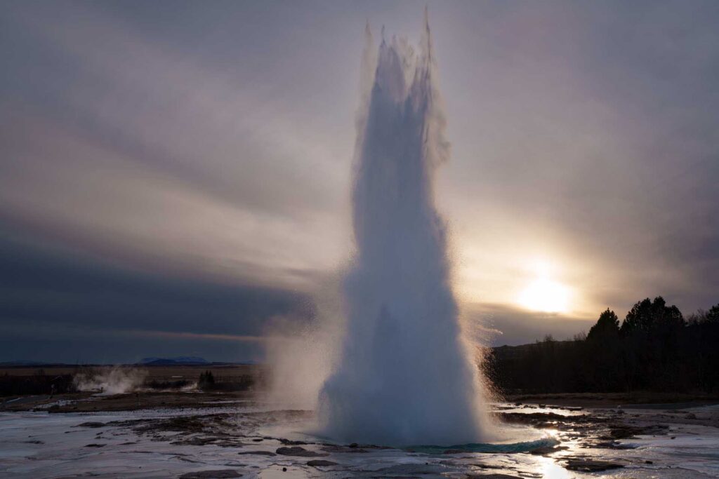 Geyser erupting in a tall column of water and steam at sunset, with dark trees and low hills on the horizon.