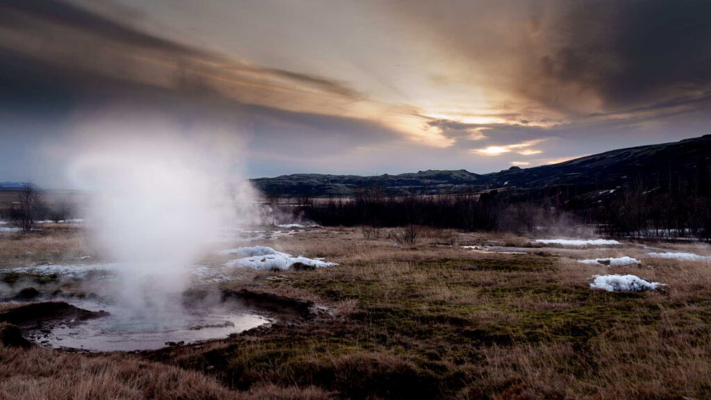 Steam billowing from a geothermal vent in a grassy field with patches of snow and hills under a dramatic evening sky.
