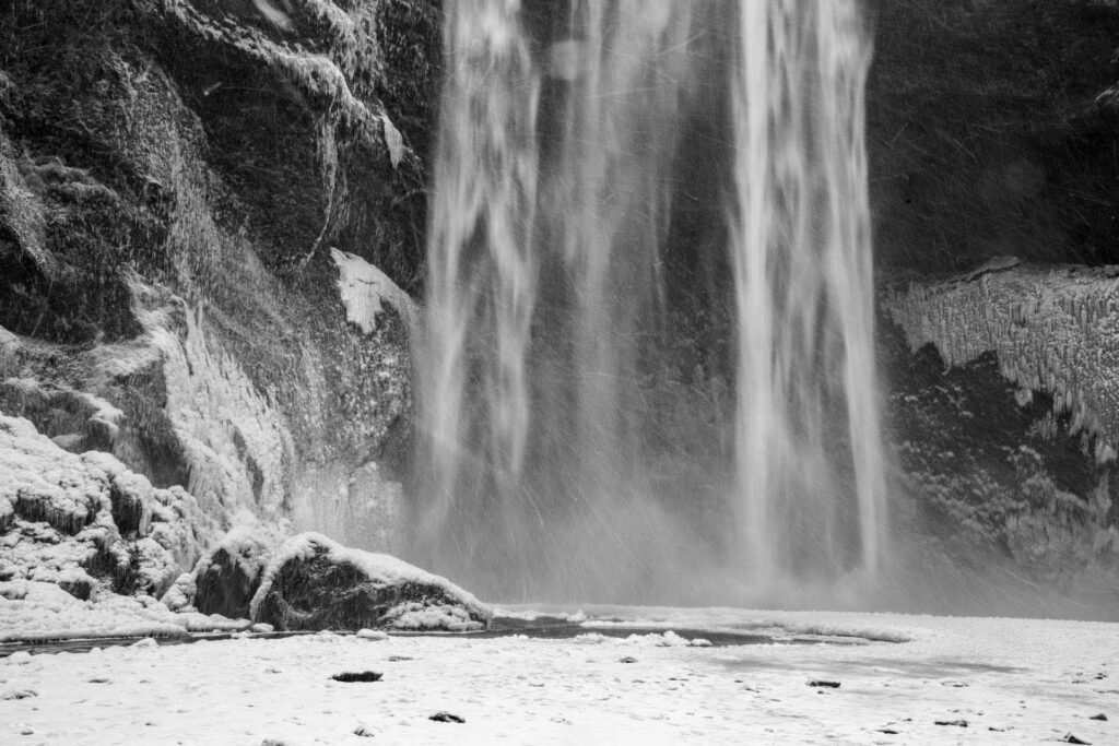 Black-and-white close-up of the base of a waterfall, showing icy rocks, drifting spray, and snow-covered ground.