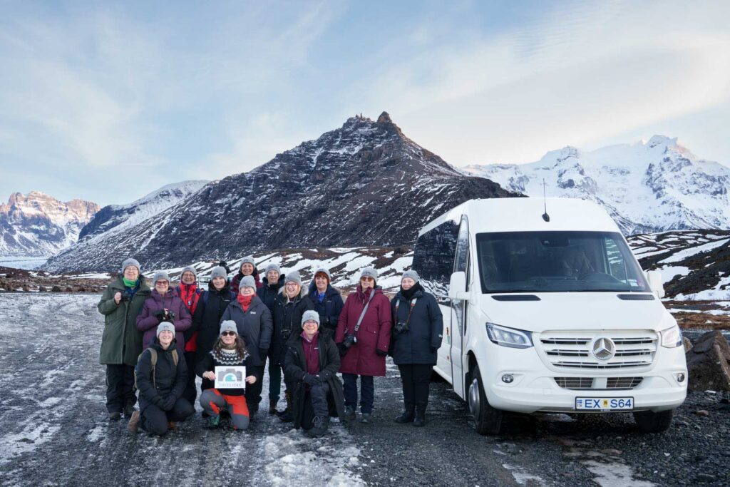 Group of bundled-up female photographers posing beside a white Mercedes van on a snowy roadside, with rugged mountains and glaciers in the background.