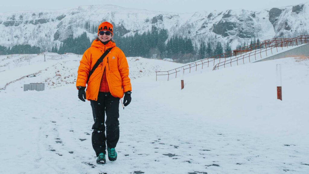 Person in a bright orange jacket and hat walking on a snowy path during light snowfall, with a rocky, tree-lined ridge in the background.
