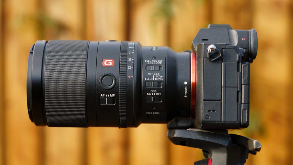 Side view of Sony FE 100mm F2.8 Macro GM OSS mounted on a Sony mirrorless camera on a tripod, showing focus ring, AF/MF switch, and OSS controls against a blurred wooden background.