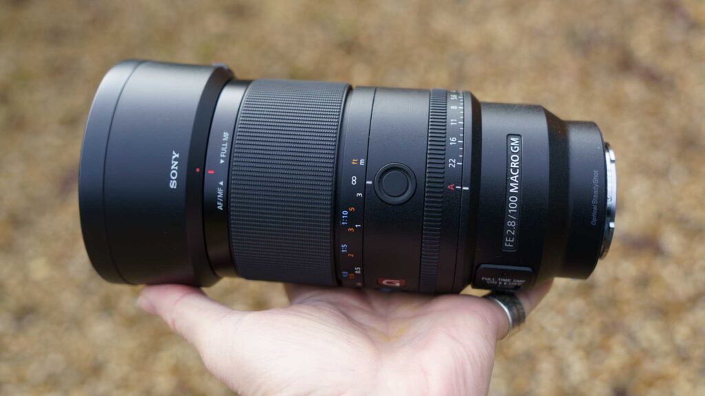 Side profile of the Sony FE 100mm F2.8 Macro GM OSS lens in hand with lens hood attached, highlighting textured focus ring and aperture markings.