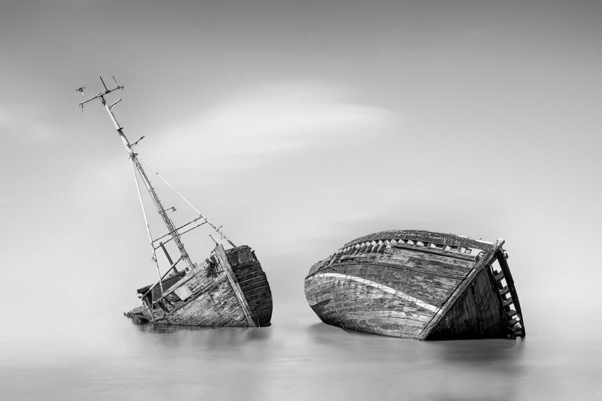 Two partially sunken wooden boats in calm water under a minimal, gray sky.