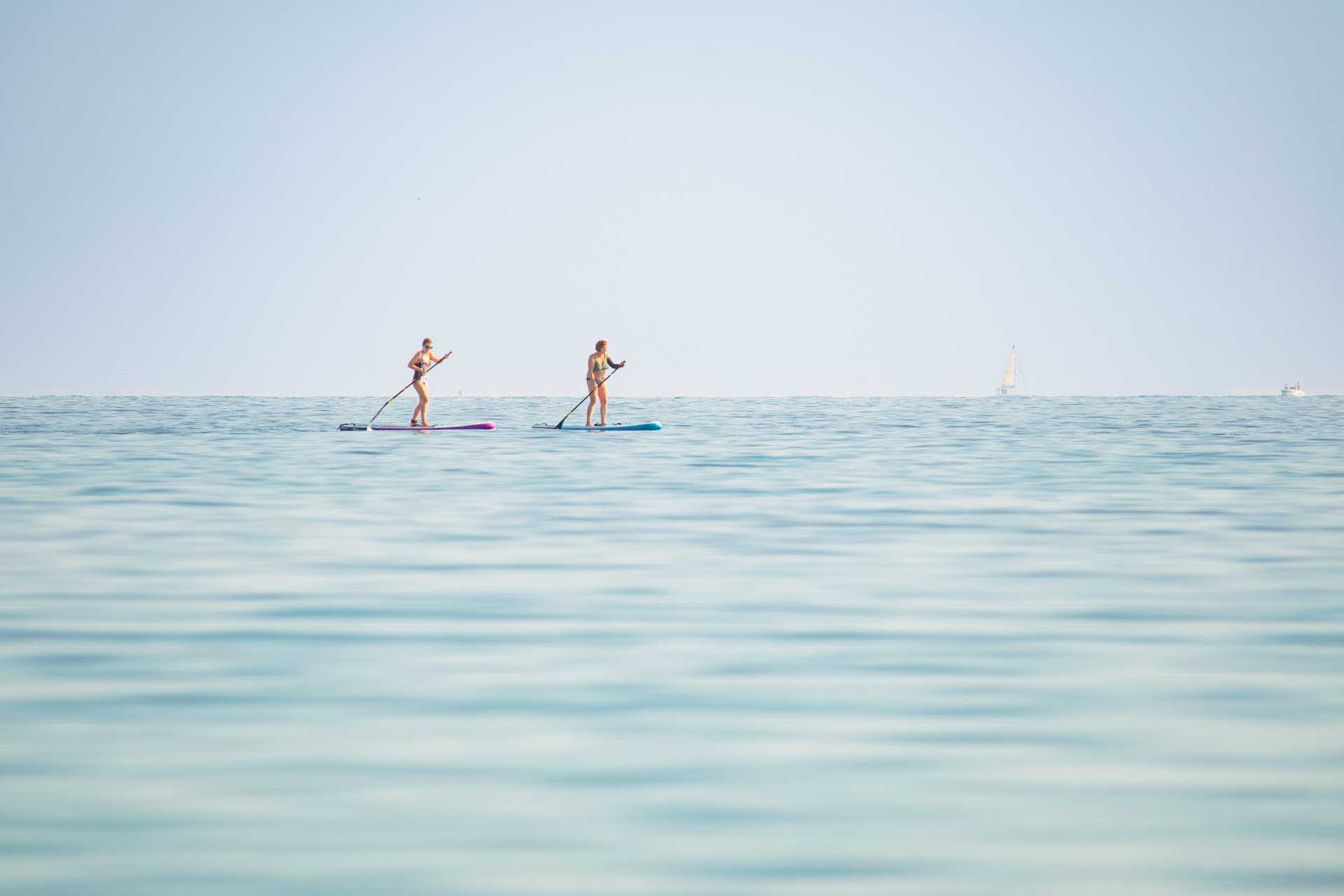 Two people paddleboarding on a calm sea with a distant sailboat on the horizon.