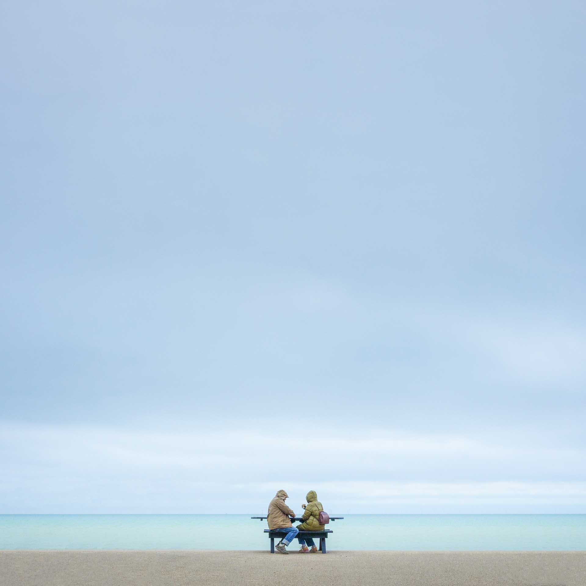 Two people sitting on a bench facing a calm sea under a wide, pale sky.