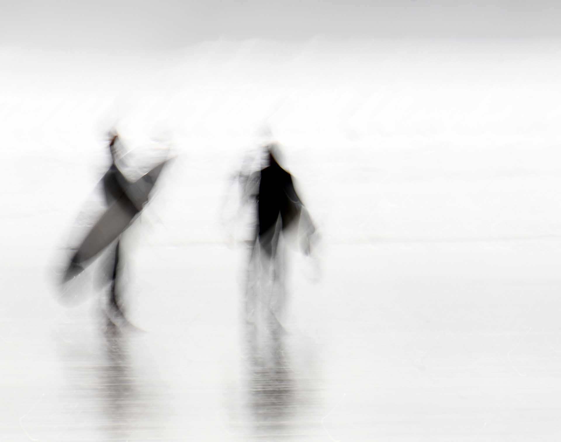 Two blurred figures walking with surfboards in a bright, minimalist beach scene.