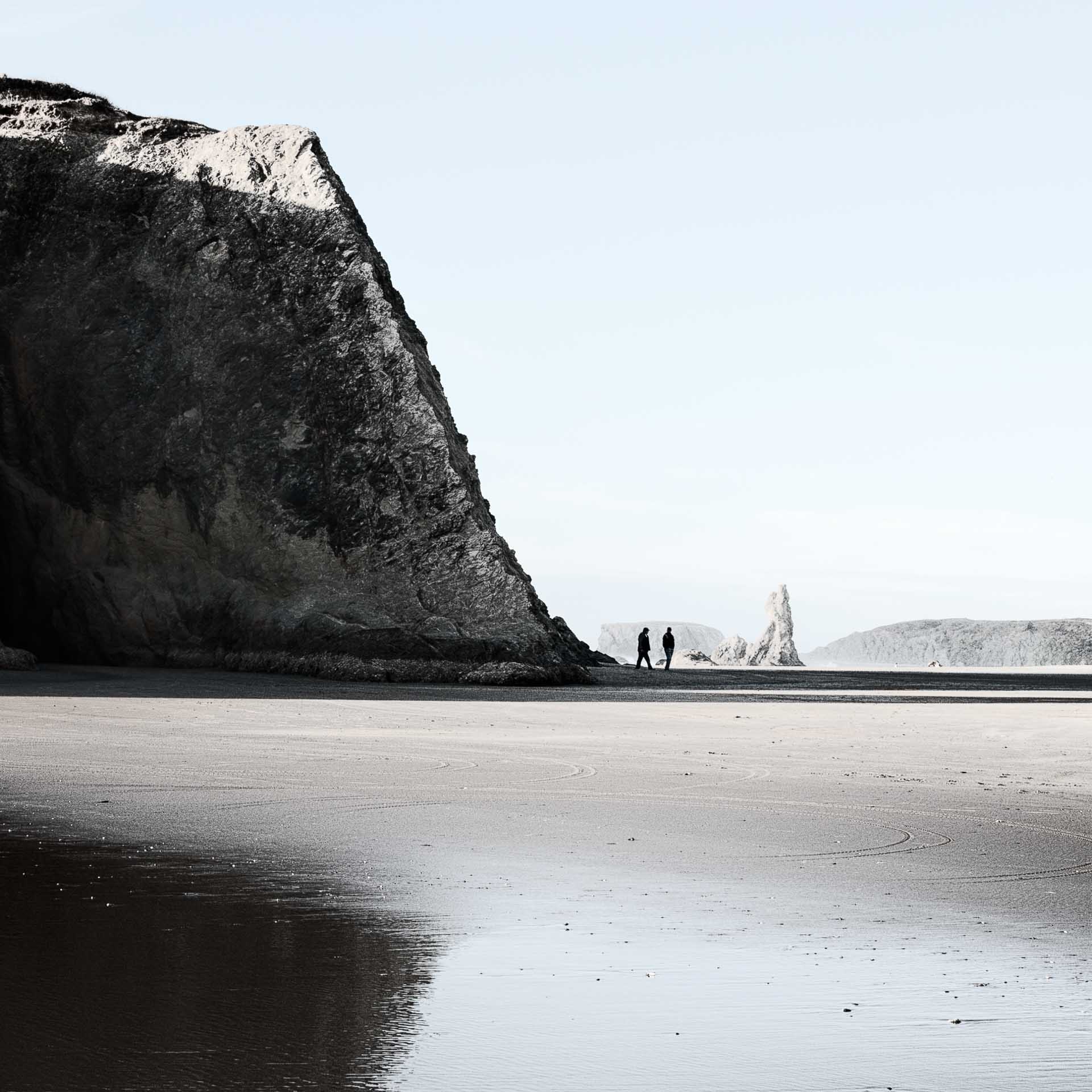 Two small figures walking along a wide, empty beach beside a large rocky cliff under a pale sky.