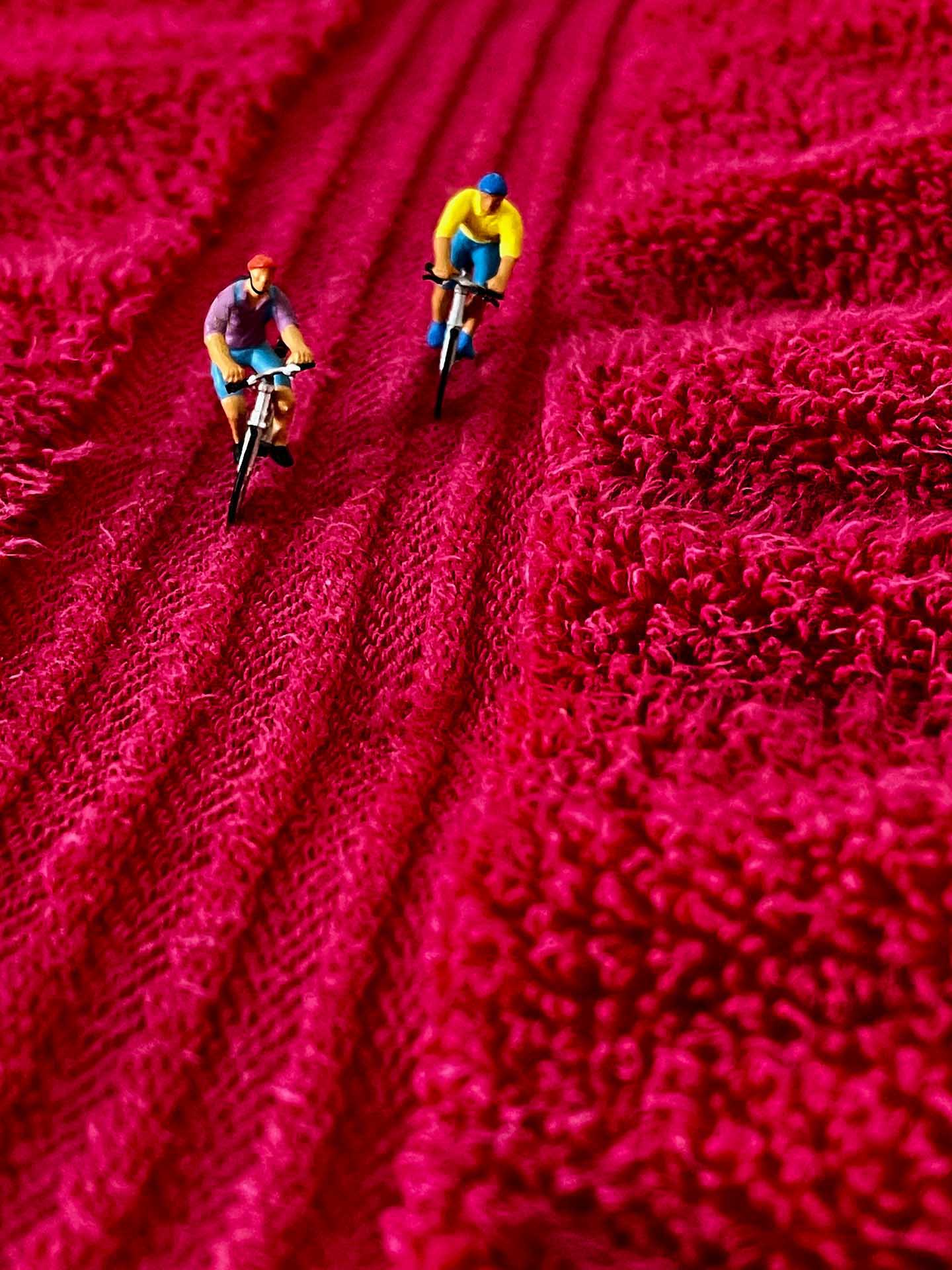Two miniature cyclists riding across the textured surface of a bright red fabric resembling rolling terrain.