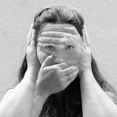 Black-and-white portrait of a woman (Mairi Clare Dunlin) with layered hands covering her eyes, ears, and mouth, leaving one eye visible through overlapping fingers.