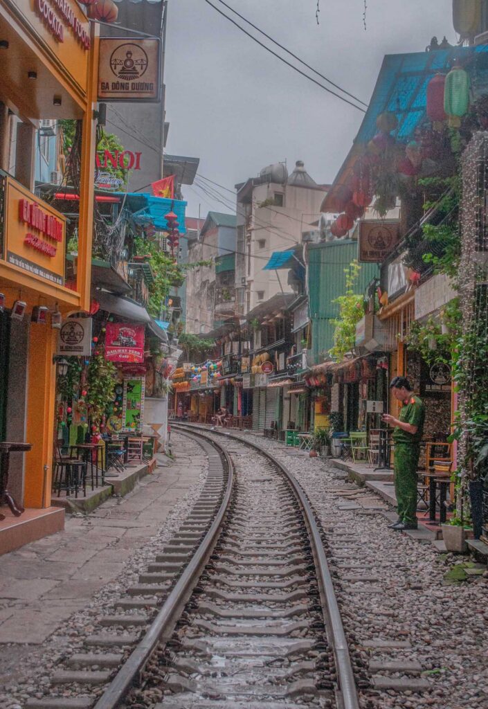 Narrow street with railway tracks running through colorful buildings and cafés; signs read “GA ĐÔNG DƯƠNG,” “HANOI,” and “Railway Train Cafe,” with a person standing beside the tracks.
