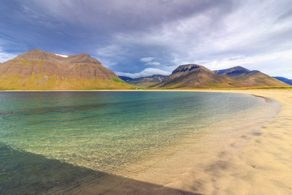 Clear shallow water along a sandy beach with footprints, curving toward distant mountains under a partly cloudy sky.