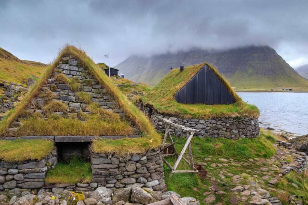 Stone turf houses with grass-covered roofs beside a rocky shoreline, with misty mountains and water in the background.