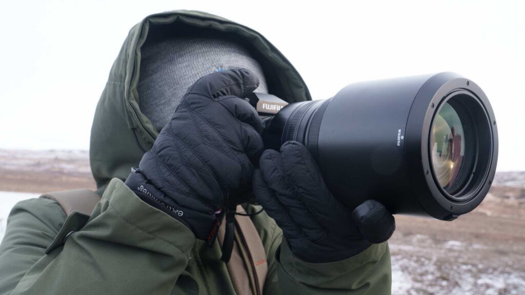 Person wearing black gloves using a Fujifilm camera with a large lens in a cold outdoor setting.