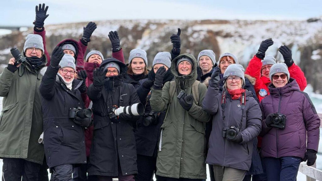Group of bundled-up photographers wearing winter jackets and grey beanies, smiling and waving at the camera in a snowy landscape.