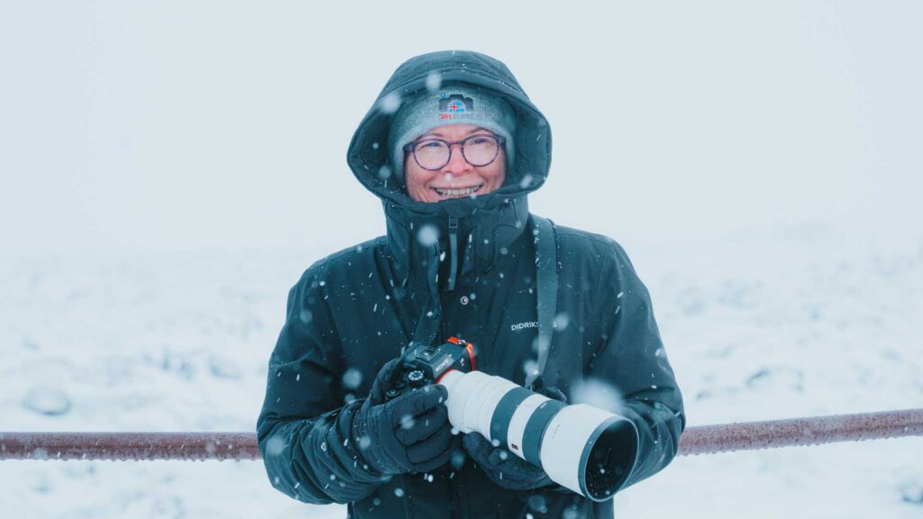 Smiling person in a hooded jacket and glasses holding a camera with a telephoto lens as snow falls around them.
