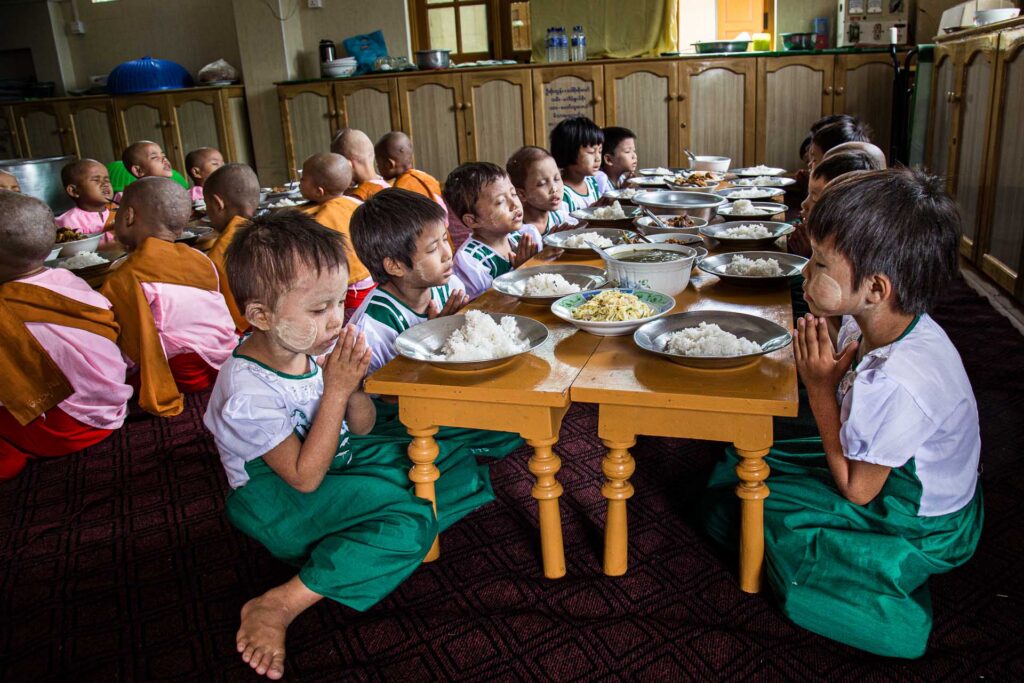 Children seated at low tables with bowls of rice, hands pressed together in prayer before a meal