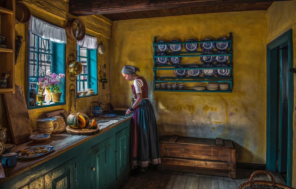 Woman in traditional clothing preparing food in a rustic kitchen with yellow walls and blue shelves of dishes