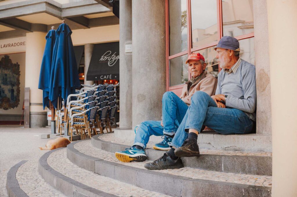 Two older men sitting on stone steps outside a café with stacked chairs and umbrellas nearby