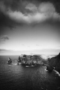 Black and white view of rugged coastal cliffs and sea stacks under heavy clouds above a dark ocean