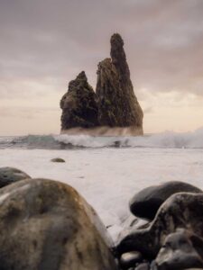 Waves crashing around a tall jagged sea stack with smooth stones in the foreground at sunset