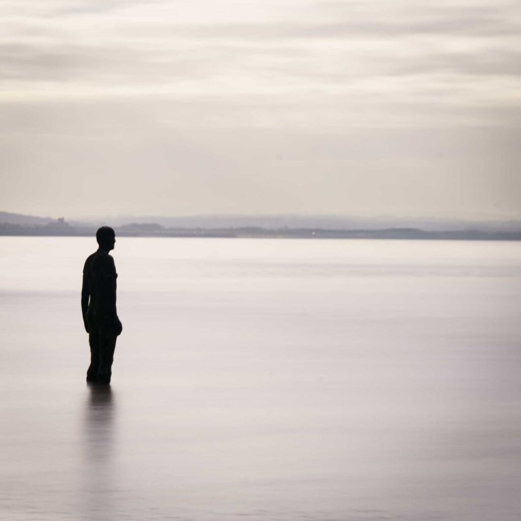 Closer view of iron figure in water, side profile against a pale horizon with low hills faintly visible across the bay