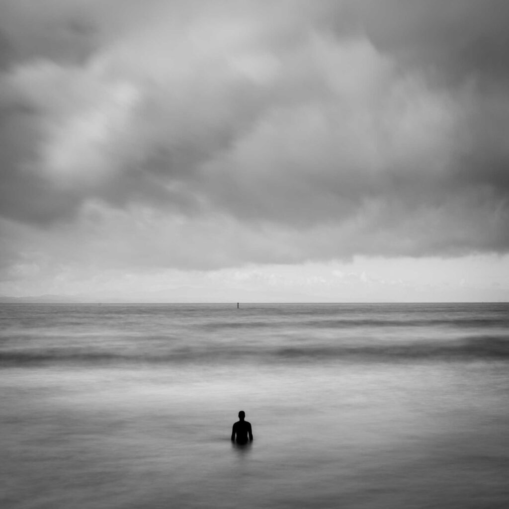 Iron figure small against wide seascape with layered waves and heavy cloud cover, horizon line stretching across the frame.
