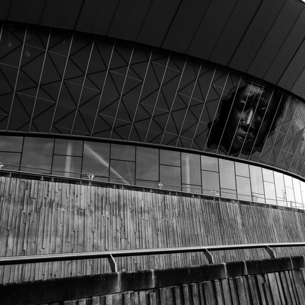 Curved modern building facade at Albert Dock with geometric glass panels and a large face artwork appearing to break through the upper wall.