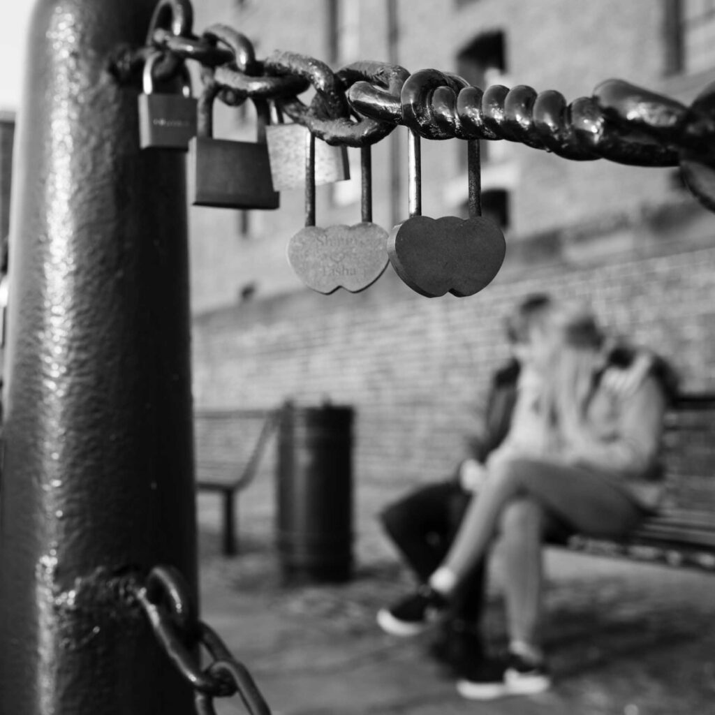 Close-up of padlocks attached to a chain at Albert Dock, with two heart-shaped locks in focus and a blurred couple sitting on a bench behind.