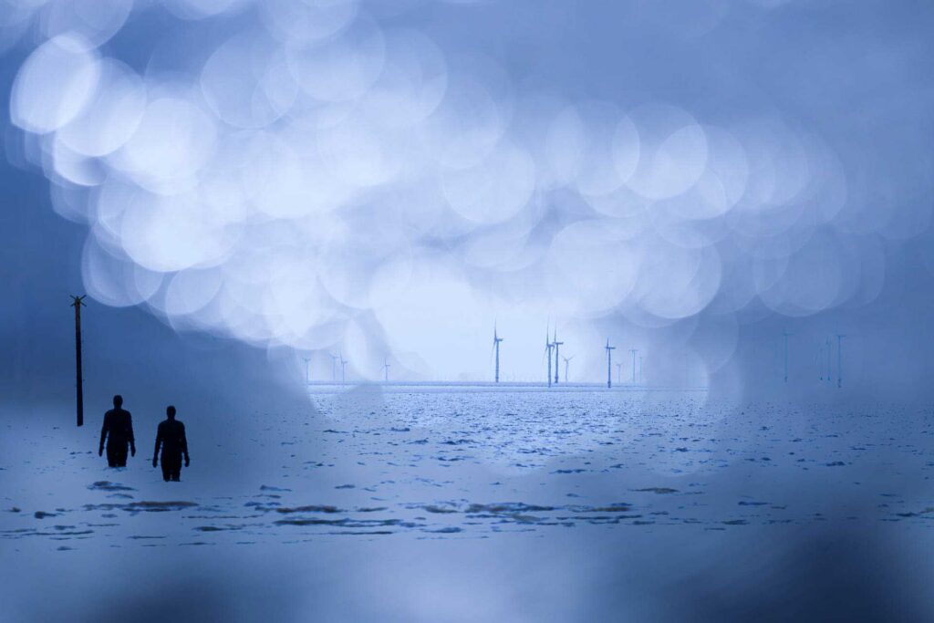 Two iron figures walking through shallow water at Crosby, overlaid with soft blue bokeh and distant wind turbines along the horizon.