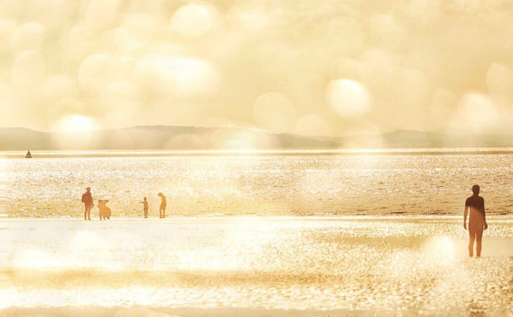 Golden-toned beach scene at Crosby with distant people and a solitary iron figure, overlaid with warm circular bokeh highlights.