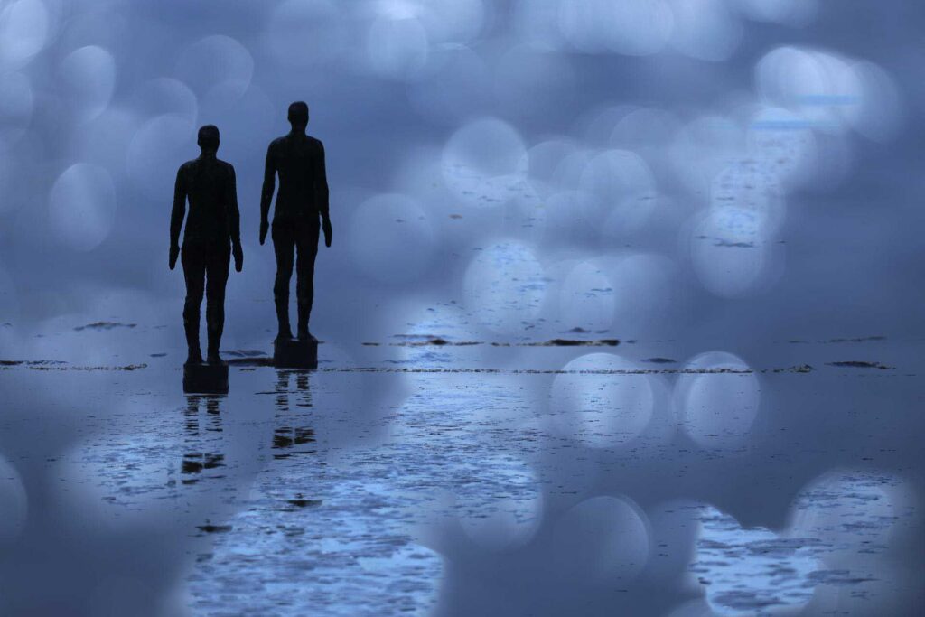 Two iron figures standing side by side in reflective water, combined with cool blue tones and large soft bokeh circles creating a dreamlike effect.