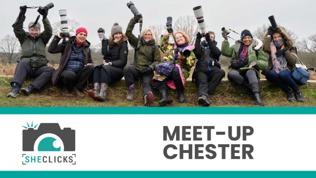 Group of women photographers seated outdoors with raised cameras; banner below shows SheClicks logo and text “MEET-UP CHESTER”.