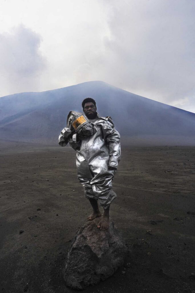 Person in reflective suit standing barefoot on a rock in a barren landscape with a volcano behind.