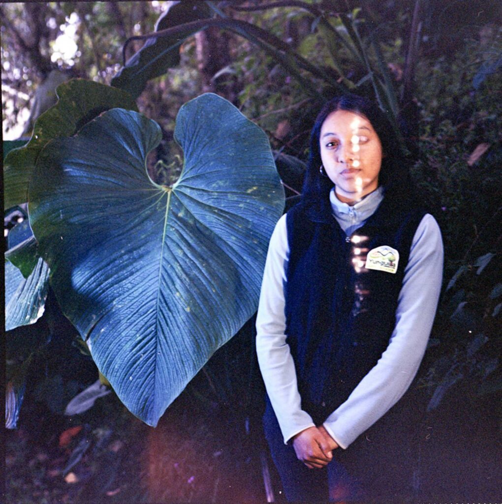 Woman standing beside a large heart-shaped leaf in a dense, dimly lit garden.