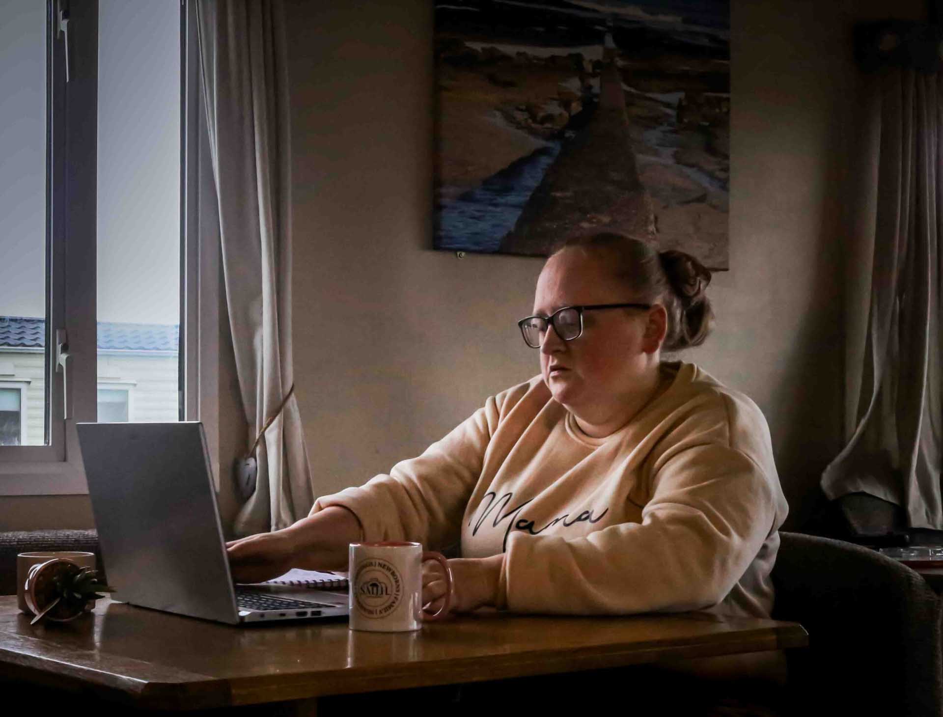 Person wearing glasses and a beige sweatshirt working on a laptop at a table beside a window, with a mug and notebook nearby.
